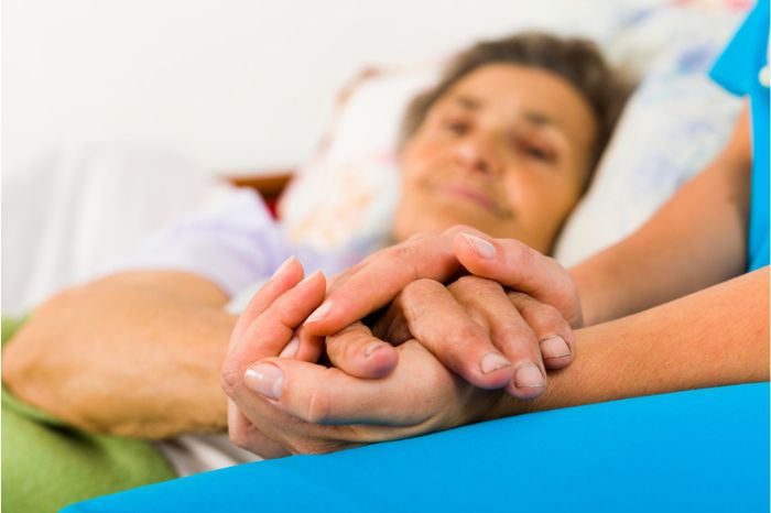 A carer holds the hand of an elderly lady lying in bed
